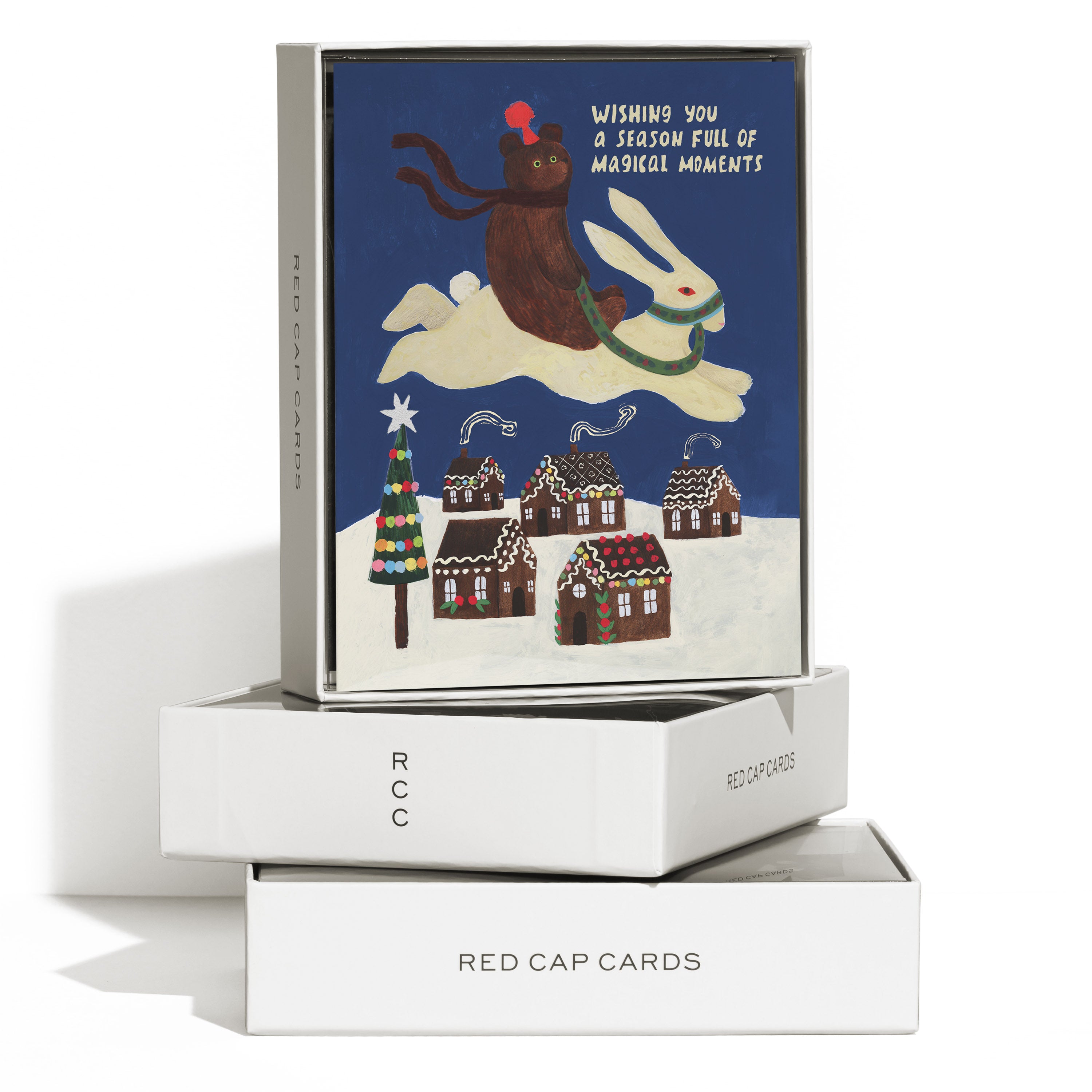 Boxed set of 8 Christmas cards with a cartoon bear riding a bunny over a snowy gingerbread town, text on blue background.